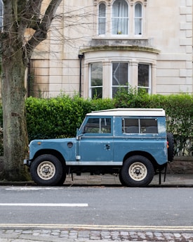 A vintage blue Land Rover is parked on the street in front of a stone building with large windows and neatly trimmed hedges. A large tree stands to the left of the vehicle, and the road is lined with cobblestones and asphalt.