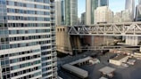 A cityscape with modern skyscrapers featuring glass and concrete facades. An elevated highway with traffic runs through the middle, including trucks with visible branding. Below the highway, there is an industrial area with buildings and parked vehicles. The image captures a mix of urban architecture and infrastructure.