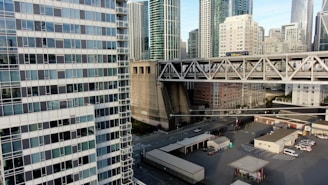 A cityscape with modern skyscrapers featuring glass and concrete facades. An elevated highway with traffic runs through the middle, including trucks with visible branding. Below the highway, there is an industrial area with buildings and parked vehicles. The image captures a mix of urban architecture and infrastructure.