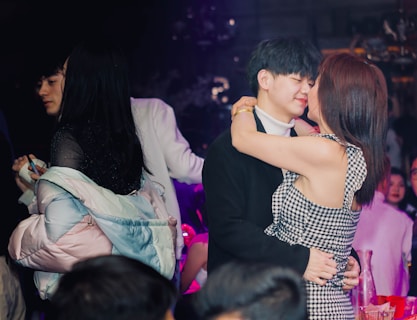 A young couple is embracing and smiling affectionately at each other in a crowded indoor setting. Behind them, another pair of individuals are interacting closely. The scene is dimly lit, with purple and blue lighting creating a party or club atmosphere. There are several people in the background, suggesting a social gathering.