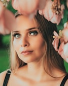 A woman with long, blonde hair gazes contemplatively at the camera. Her eyes are striking, accentuated by long eyelashes. Pink flowers in the foreground frame her face, adding a soft, dreamy quality. The background is blurred greenery, suggesting an outdoor setting.