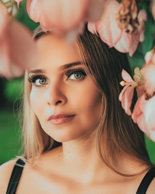 A woman with long, blonde hair gazes contemplatively at the camera. Her eyes are striking, accentuated by long eyelashes. Pink flowers in the foreground frame her face, adding a soft, dreamy quality. The background is blurred greenery, suggesting an outdoor setting.