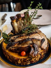 A succulent, well-cooked piece of meat with bones, garnished with fresh herbs like rosemary and thyme, presented on a rustic wooden board. A small red cherry tomato and green leaves are placed beside the meat, creating an attractive and appetizing presentation. The background includes a white tablecloth and a few plates and cutlery.
