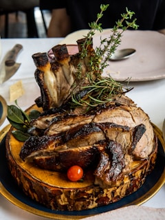 A succulent, well-cooked piece of meat with bones, garnished with fresh herbs like rosemary and thyme, presented on a rustic wooden board. A small red cherry tomato and green leaves are placed beside the meat, creating an attractive and appetizing presentation. The background includes a white tablecloth and a few plates and cutlery.