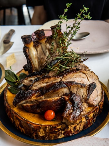 A succulent, well-cooked piece of meat with bones, garnished with fresh herbs like rosemary and thyme, presented on a rustic wooden board. A small red cherry tomato and green leaves are placed beside the meat, creating an attractive and appetizing presentation. The background includes a white tablecloth and a few plates and cutlery.
