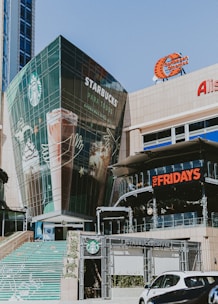 A modern commercial building featuring large glass windows, prominently displaying logos for Starbucks and TGI Fridays. The area includes outdoor signage for a Caribbean Cinemas and other shops. A set of stairs decorated with colorful designs leads up to the building entrance.
