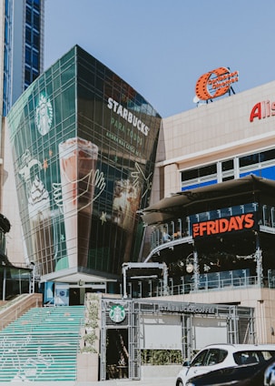 A modern commercial building featuring large glass windows, prominently displaying logos for Starbucks and TGI Fridays. The area includes outdoor signage for a Caribbean Cinemas and other shops. A set of stairs decorated with colorful designs leads up to the building entrance.