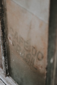 A weathered, vintage coffee sack is visible through a dusty window, partially obscured by the frame. The faded text 'CAFÉ DO BRASIL' is faintly printed on the burlap material.