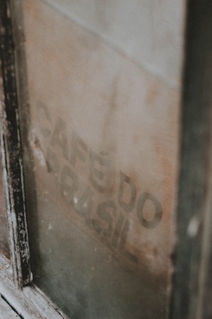 A weathered, vintage coffee sack is visible through a dusty window, partially obscured by the frame. The faded text 'CAFÉ DO BRASIL' is faintly printed on the burlap material.