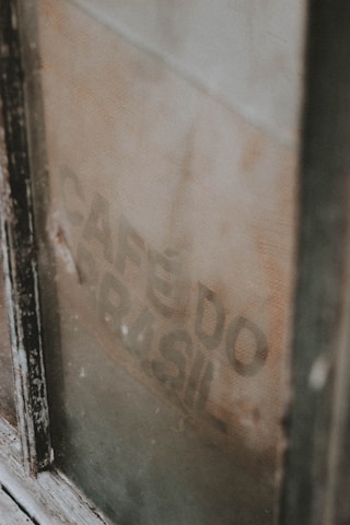 A weathered, vintage coffee sack is visible through a dusty window, partially obscured by the frame. The faded text 'CAFÉ DO BRASIL' is faintly printed on the burlap material.