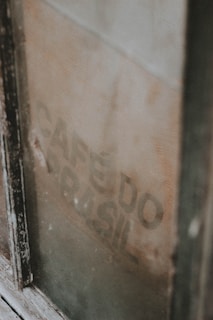 A weathered, vintage coffee sack is visible through a dusty window, partially obscured by the frame. The faded text 'CAF&Eacute; DO BRASIL' is faintly printed on the burlap material.