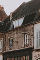 An old building with weathered walls and a sign that reads 'PHOTOGRAPHIE'. The structure has a sloped roof with dark, aged tiles and multiple windows. A street lamp is attached to the front of the building. The building appears to be part of a quaint, old-fashioned neighborhood.