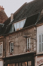 An old building with weathered walls and a sign that reads 'PHOTOGRAPHIE'. The structure has a sloped roof with dark, aged tiles and multiple windows. A street lamp is attached to the front of the building. The building appears to be part of a quaint, old-fashioned neighborhood.