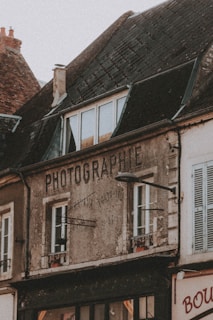 An old building with weathered walls and a sign that reads 'PHOTOGRAPHIE'. The structure has a sloped roof with dark, aged tiles and multiple windows. A street lamp is attached to the front of the building. The building appears to be part of a quaint, old-fashioned neighborhood.