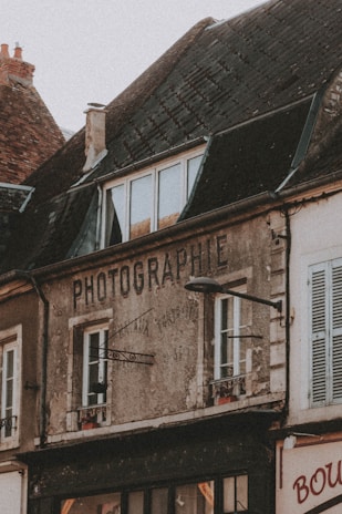 An old building with weathered walls and a sign that reads 'PHOTOGRAPHIE'. The structure has a sloped roof with dark, aged tiles and multiple windows. A street lamp is attached to the front of the building. The building appears to be part of a quaint, old-fashioned neighborhood.