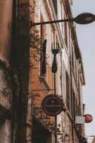 A rustic outdoor cafe sign hangs on the side of a weathered building along a narrow European street. The sign, stylized with a fork and knife, advertises DolceVita cafe. Climbing vines and warm sunlight add a nostalgic charm to the scene.