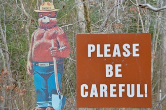 A cartoon figure of a bear wearing a ranger hat and pants stands next to a large brown sign with bold white letters that read 'PLEASE BE CAREFUL'. The bear is holding a shovel and has a serious expression.