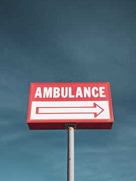 A red sign with the word 'Ambulance' in white uppercase letters is mounted on a pole against a clear blue sky. An arrow beneath the text points in one direction.