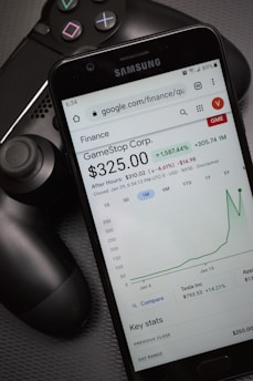 A smartphone displays a Google Finance page showing stock information for GameStop Corp., with a graph indicating a significant rise in stock price. The phone screen prominently shows a price of $325.00. Next to the phone is a black gaming controller, suggesting a connection to video gaming.