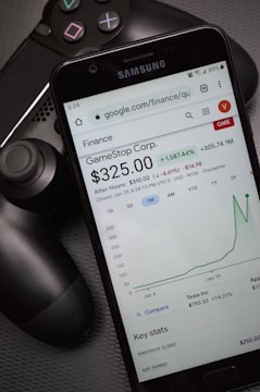 A smartphone displays a Google Finance page showing stock information for GameStop Corp., with a graph indicating a significant rise in stock price. The phone screen prominently shows a price of $325.00. Next to the phone is a black gaming controller, suggesting a connection to video gaming.