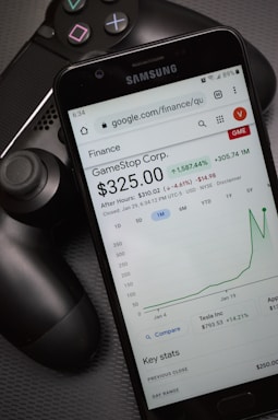 A smartphone displays a Google Finance page showing stock information for GameStop Corp., with a graph indicating a significant rise in stock price. The phone screen prominently shows a price of $325.00. Next to the phone is a black gaming controller, suggesting a connection to video gaming.
