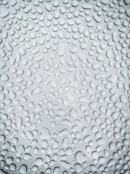 An array of water droplets of varying sizes covers a smooth surface, creating a textured pattern. The light reflection on the droplets gives them a glossy appearance and enhances the depth of the image.