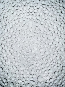 An array of water droplets of varying sizes covers a smooth surface, creating a textured pattern. The light reflection on the droplets gives them a glossy appearance and enhances the depth of the image.