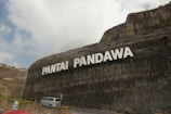 A large stone cliff displays the words 'Pantai Pandawa' in bold white letters. Below, a road curves along the base of the cliff with a silver car parked near a stone wall. Some greenery is visible at the top and sides of the cliff. The sky is partly cloudy, casting soft shadows on the scene.