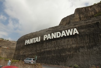 A large stone cliff displays the words 'Pantai Pandawa' in bold white letters. Below, a road curves along the base of the cliff with a silver car parked near a stone wall. Some greenery is visible at the top and sides of the cliff. The sky is partly cloudy, casting soft shadows on the scene.