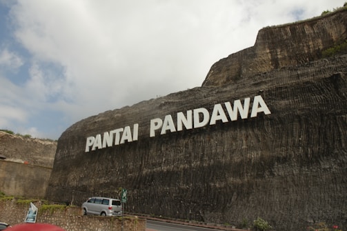 A large stone cliff displays the words 'Pantai Pandawa' in bold white letters. Below, a road curves along the base of the cliff with a silver car parked near a stone wall. Some greenery is visible at the top and sides of the cliff. The sky is partly cloudy, casting soft shadows on the scene.