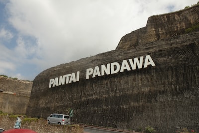 A large stone cliff displays the words 'Pantai Pandawa' in bold white letters. Below, a road curves along the base of the cliff with a silver car parked near a stone wall. Some greenery is visible at the top and sides of the cliff. The sky is partly cloudy, casting soft shadows on the scene.