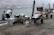 Two individuals are unloading or organizing a variety of boxes and crates containing goods, possibly fruits or vegetables, beside a canal. The atmosphere is misty, partially obscuring the background and giving a hint of old Venetian buildings across the water. Several hand carts filled with boxes are positioned nearby on a cobblestone path.