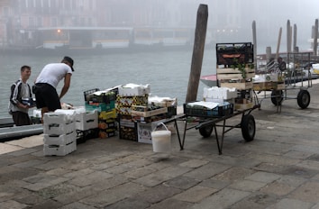 Two individuals are unloading or organizing a variety of boxes and crates containing goods, possibly fruits or vegetables, beside a canal. The atmosphere is misty, partially obscuring the background and giving a hint of old Venetian buildings across the water. Several hand carts filled with boxes are positioned nearby on a cobblestone path.
