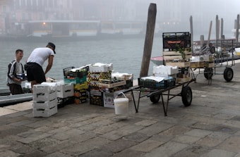 Two individuals are unloading or organizing a variety of boxes and crates containing goods, possibly fruits or vegetables, beside a canal. The atmosphere is misty, partially obscuring the background and giving a hint of old Venetian buildings across the water. Several hand carts filled with boxes are positioned nearby on a cobblestone path.