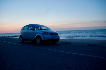 A silver car is parked on a coastal road during sunset, with a serene ocean view in the background. The sky transitions from soft orange and pink hues near the horizon to a deepening blue overhead.