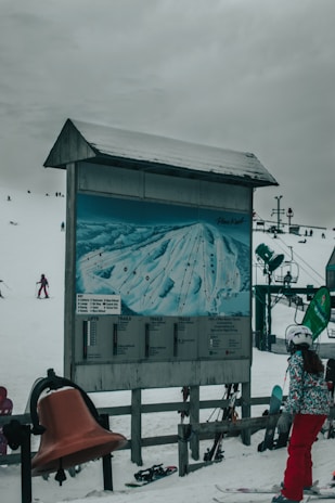A snowy ski resort with a large trail map displayed on a board. Skiers are visible in the background, and a skier stands near the board looking at the map. The sky is overcast, and ski equipment can be seen scattered around.