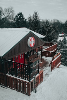 A wooden cabin with black railings and a red first aid sign is situated in a snowy landscape. The cabin is surrounded by evergreen trees, and snow covers the ground and roof. The atmosphere appears cold and wintry.