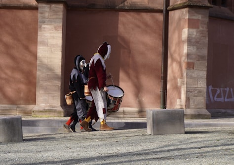 Two individuals in medieval jester costumes are seen walking along a cobblestone path. They carry traditional drums, suggesting a performance or parade setting. The background is a historic brick wall with columns, and the scene is set in daylight.