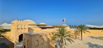 A traditional Middle Eastern architectural structure with arched windows and earthy tones, set against a clear blue sky. Palm trees are positioned in the courtyard, and a red flag is flying on a tall pole.
