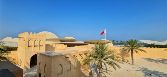 A traditional Middle Eastern architectural structure with arched windows and earthy tones, set against a clear blue sky. Palm trees are positioned in the courtyard, and a red flag is flying on a tall pole.