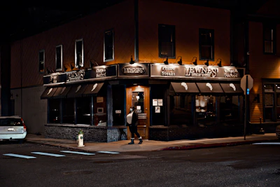 A warmly lit steakhouse on a city corner during the night, featuring dark awnings and prominent signage. A person is walking towards the entrance, which is illuminated and inviting. A car is parked on the side street, and there is a fire hydrant nearby.