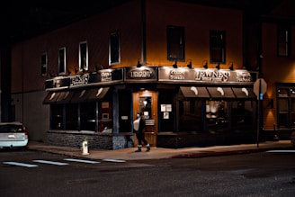 A warmly lit steakhouse on a city corner during the night, featuring dark awnings and prominent signage. A person is walking towards the entrance, which is illuminated and inviting. A car is parked on the side street, and there is a fire hydrant nearby.