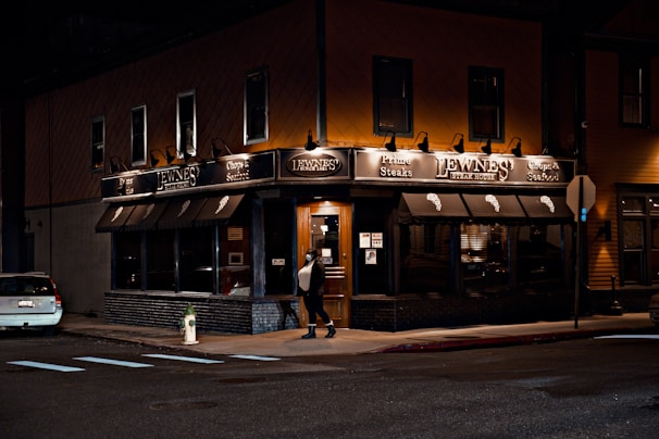 A warmly lit steakhouse on a city corner during the night, featuring dark awnings and prominent signage. A person is walking towards the entrance, which is illuminated and inviting. A car is parked on the side street, and there is a fire hydrant nearby.