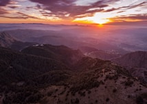 A breathtaking landscape of mountainous terrain at sunset with layers of hills and valleys extending into the distance. The sky is colored in hues of purple, pink, and orange as the sun descends, casting warm light over the rugged scenery below.