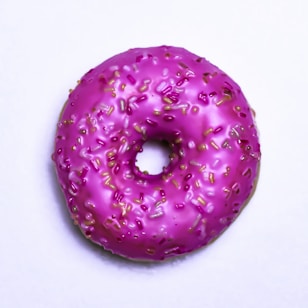 A single donut with bright pink icing and colorful sprinkles is placed on a plain white background. The donut's surface is glossy, suggesting it is fresh and possibly sweet.