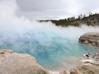 A geothermal hot spring emits thick steam that rises from its surface, creating a misty atmosphere. The water appears blue, surrounded by rocky edges and sparse vegetation. In the background, a forest with evergreen trees is visible under a cloudy sky.