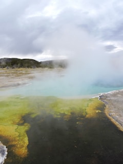 A geothermal area with a hot spring emitting steam. The water in the hot spring displays vibrant colors, primarily yellow, green, and blue. Surrounding the hot spring is a rocky terrain with sparse vegetation visible in the distance under a cloudy sky.