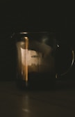 A dark, moody scene featuring a transparent glass measuring cup partially filled with liquid. The cup is marked with measurement lines from 100 to 500 milliliters. The surrounding environment is dimly lit, creating a dramatic and contemplative atmosphere.