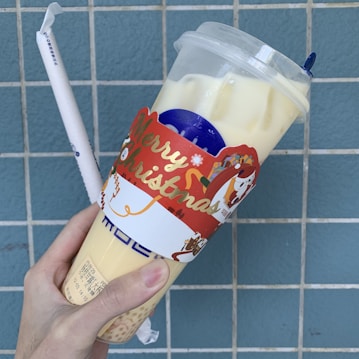 A hand holds a bubble tea cup adorned with a festive Merry Christmas sleeve. The cup is filled with a light-colored beverage and sealed with a plastic lid. In the background, there is a tiled wall in shades of blue, and a white straw with text on it is also visible.