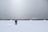 A metal bollard surrounded by fresh snow, casting a shadow on the white surface. The scene is split horizontally with a contrasting textured ground, possibly ice or pavement, above the snow.